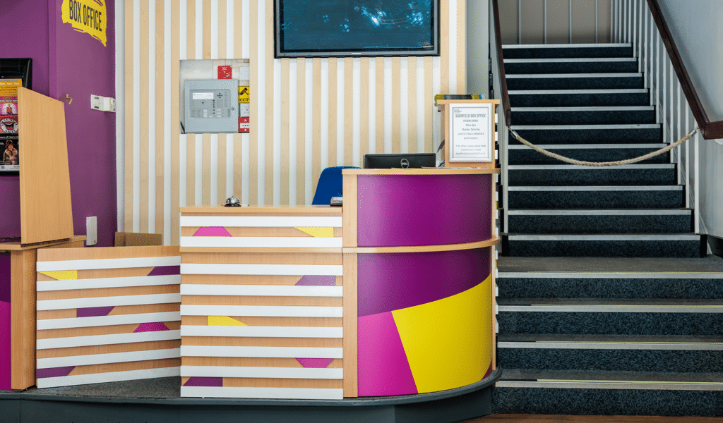The Barnfield Theatre box office. A large desk with a black top and striped wooden panelling separated with pink, purple and yellow colour blocks. On the right-hand side of the desk, a curved piece of wood with a pink, purple and yellow block overlay. Behind the desk, a desktop PCs. On the wall behind the desk, white and bare wood striped panelling. To the right-hand side of the desk area, grey carpeted stairs leading up to the Barnfield Theatre auditorium.