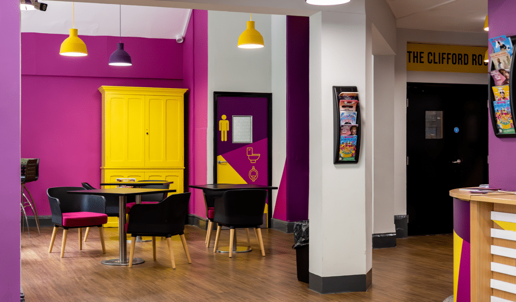 The Barnfield Theatre break area. Background: Pink and white colour walls. Pueple and yellow coloured lampshades suspended from the ceiling. Foreground: A large, yellow book cupboard. Black chairs with pink cushions sat around black surface tables. To the right-hand side of the photo, a black door with a yellow sign above it reading 'The Clifford Room'.