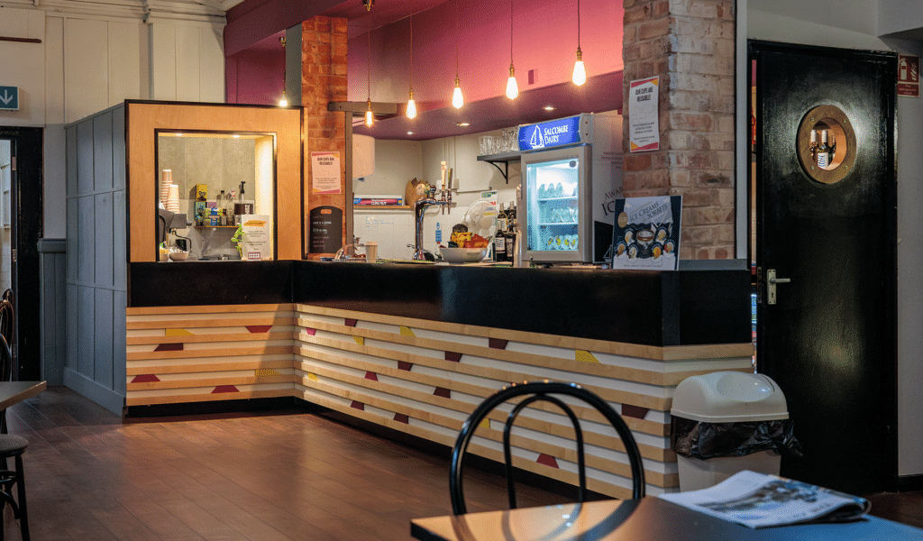 The Barnfield Theatre cafe. A long, black-surfaced bar, above striped wooden panelling separated with pink, purple and yellow colour blocks. The bar is in front of two brick pillars. Behind the bar, there is a coffee machine and ice cream fridge. Lit lightbulbs hand from the ceiling above the bar.