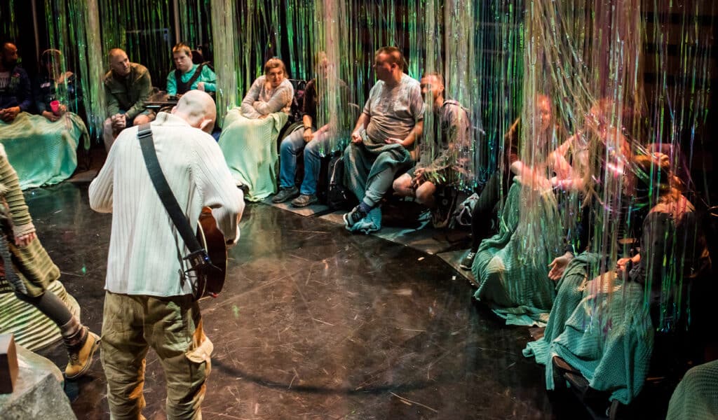 Production photo from Isle of Brimsker. A mixed group of people sit in a semi circle around a man wearing a grey jumper and brown trousers who is playing a guitar. Almost all of the people sat in the semi-circle are covered long strands of tinsel suspended from the ceiling, and many of them also wearing knitted green blankets over their legs.