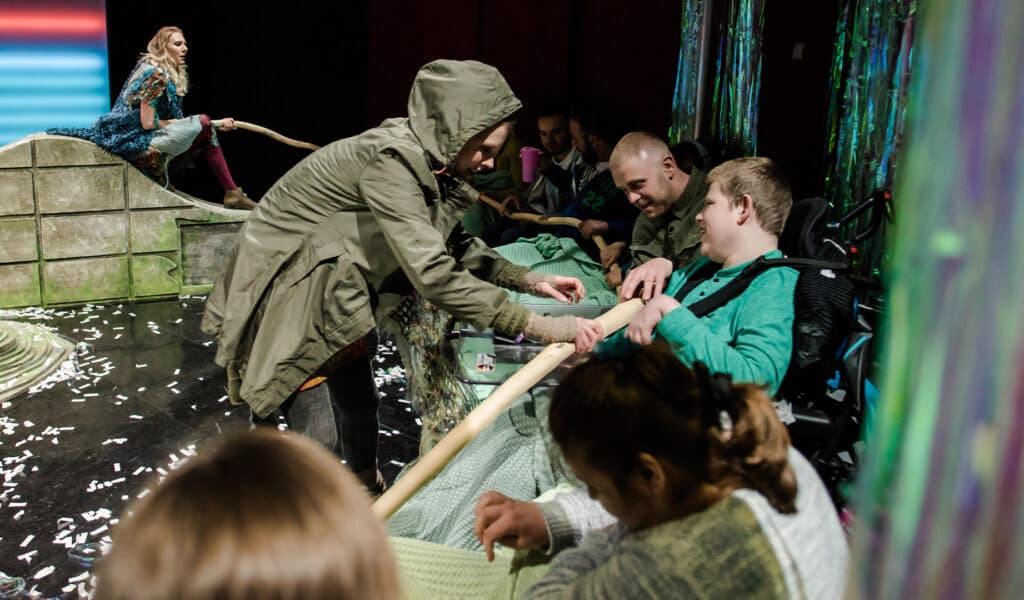 Production photo from Isle of Brimsker. A woman wearing a brown rain coat lets an audience member wearing a light green jumper using a wheelchair touch a long piece of wood. They are both smiling. They are surrounded by a mixed group of people sat in a semi-circle around a small set that is designed to look like weathered rocks. White confetti is spread across the floor.