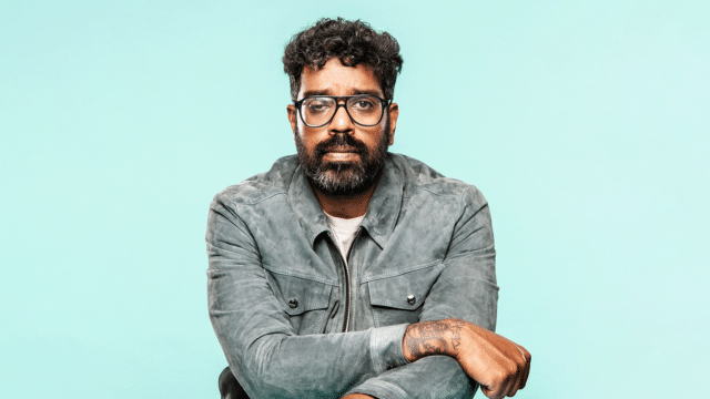 Romesh Ranganathan, wearing a grey denim jacket over a white T-shirt, sits frowning with their arms crossed in front of a light blue background.