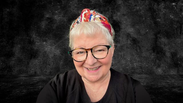 A photo of Janey Godley wearing a black t-shirt, glasses and a red bandana headband. Se smiles and looks at the camera. The background is black and textured.