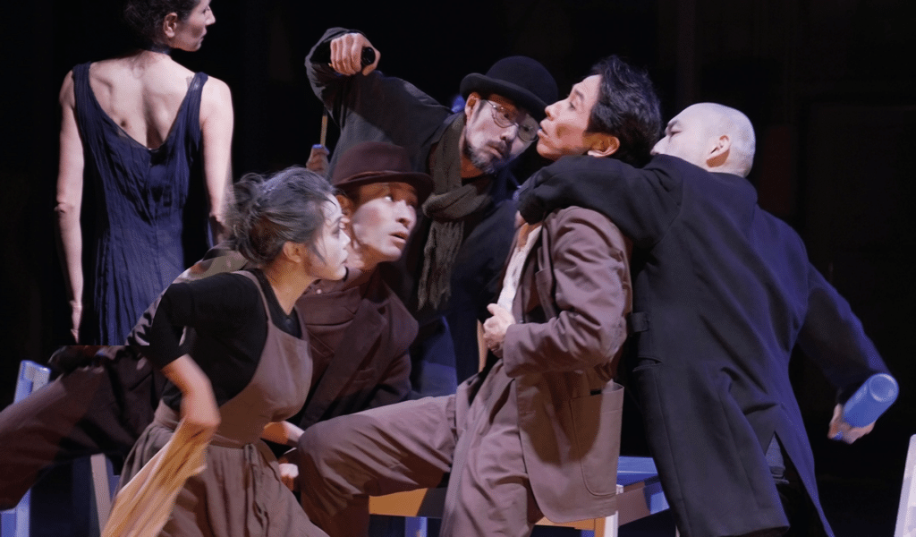 tarinainanika's Rey Camoy production photo. A mixed group of people crowd around a Japanese man, wearing a brown suit. A man wearing a black suit jacket is restraining the Japanese man, and another man is about to punch them.