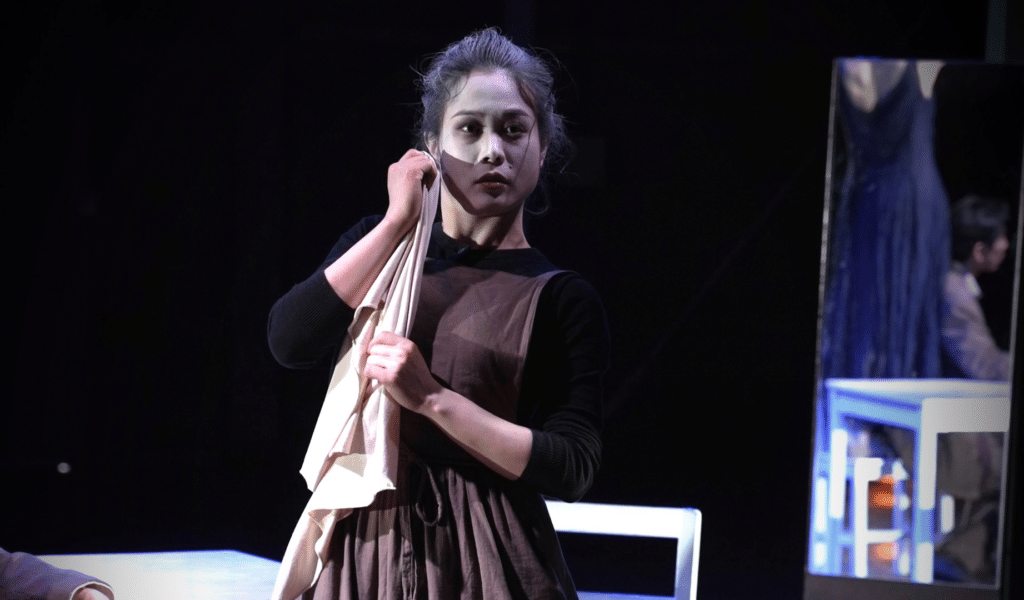 tarinainanika's Rey Camoy production photo. A young Japanese woman, wearing a brown apron over a black top, holds a cleaning rag up to her face. Behind her, a mirror, reflecting a person wearing a blue dress and a man in a grey suit sitting beside a white table.