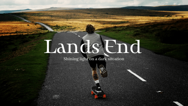 Land's End: Shining Light on a dark situation. A dark-haired man is skateboarding down a paved road, flowing hills of grass and moorland around him.