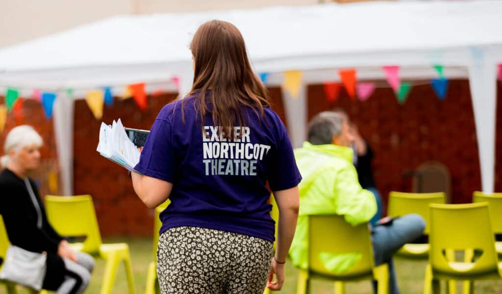 A photograph of a woman with shoulder-length brown hair. She faces away from the camera, where a marquee and green chairs can be seen. She is wearing a purple t-shirt with the Exeter Northcott Theatre logo on the back.