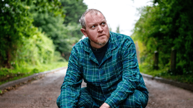 Miles Jupp: On I Bang Promo Image: Miles Jupp, wearing turquoise chequered pyjamas, crouches on a country road, frowning.