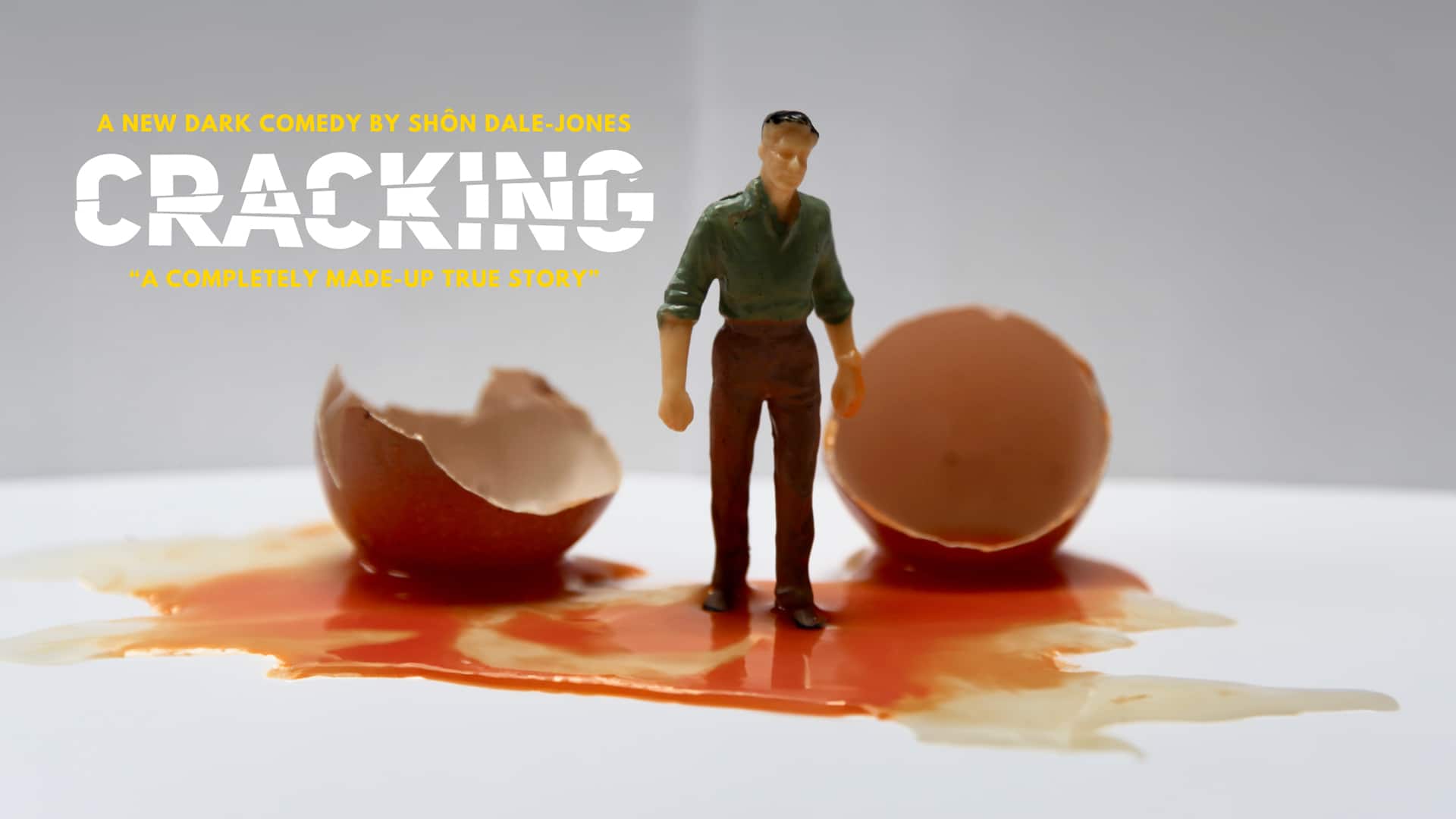 Cracking artwork. A small figurine of a man wearing a green shirt and brown trousers steps through the spilled yolk of a cracked egg on a white table, with split egg shells either side. Text reads: 'A new dark comedy by Shôn Dale-Jones. CRACKING. A completely made-up true story'.