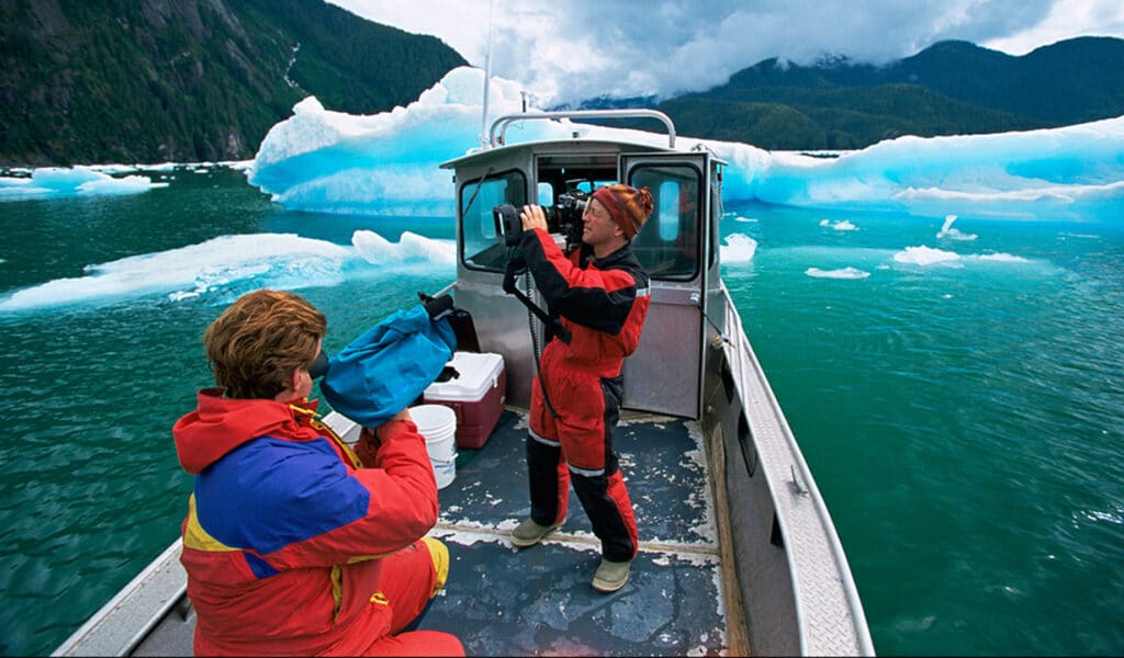 Doug Allan (centre), pictured here as a middle aged man wearing a black and red insulated jumpsuit, operates a camera while on a small boat with a middle aged woman (centre-left), wearing a blue and red insulated jumpsuit and operating another camera, as they sail through an iceberg-filled fjord.