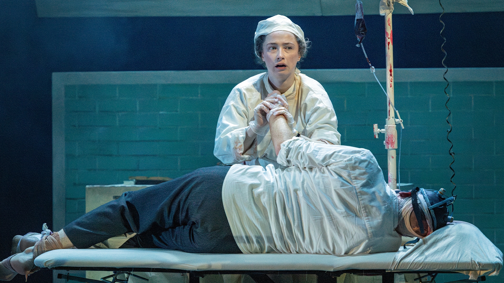 Frankenstein production photo. Theatre stage. Inside a clinical hospital room with light blue brick walls. A young woman wearing a surgeons cap and gown holds the hand of Frankenstein's monster, who is lying on a makeshift hospital bed.