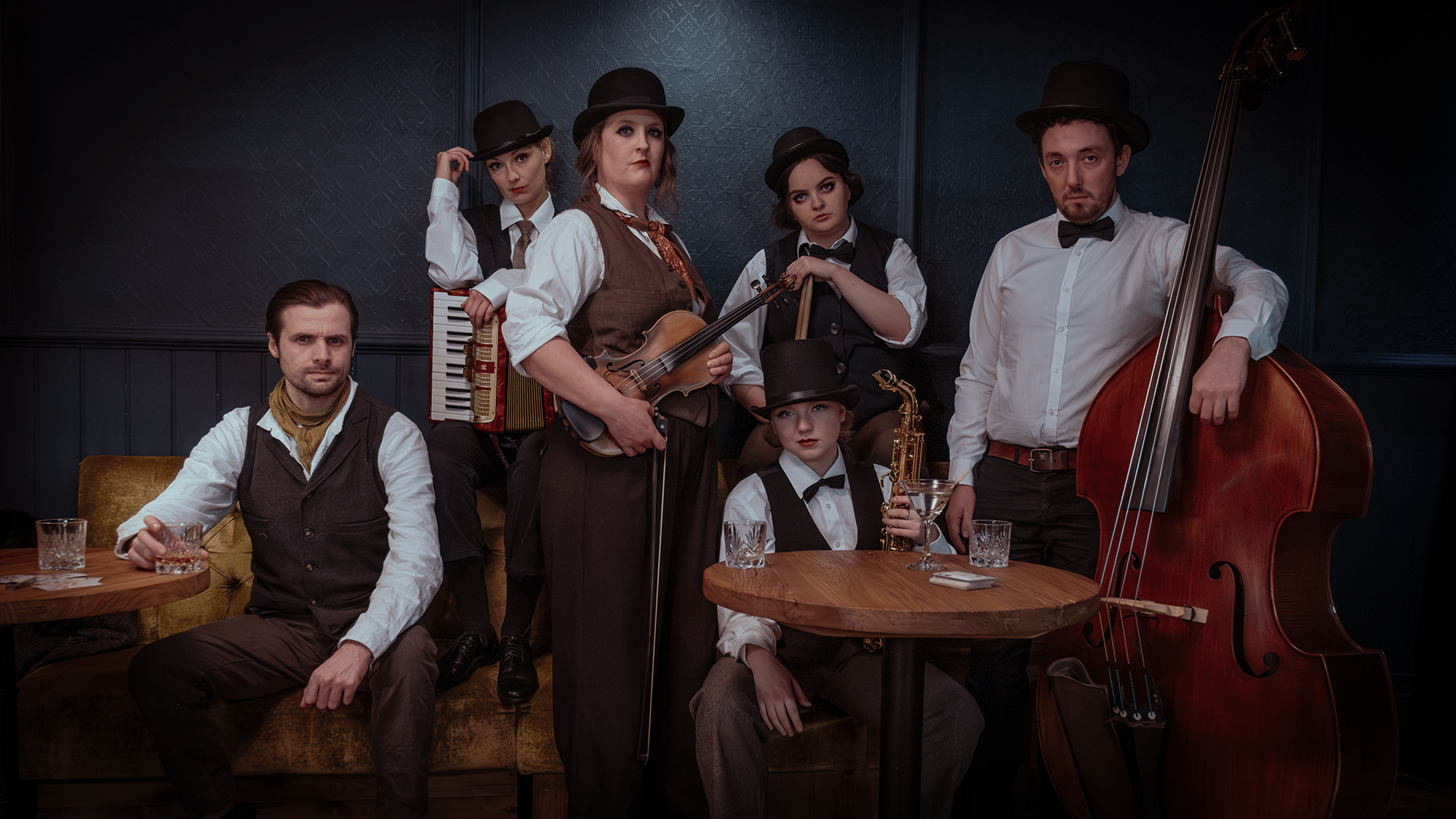 The cast of 40 Elephants as the 1920s' 40 Elephant Gang, looking cool and staring into the camera daringly, holding their instruments. Left to right: arm resting on the table, drinking his glass of whiskey; elbow on her accordeon tipping her hat; holding her violin and bow; holding drumsticks, holding her saxophone, leaning on his contrabass.