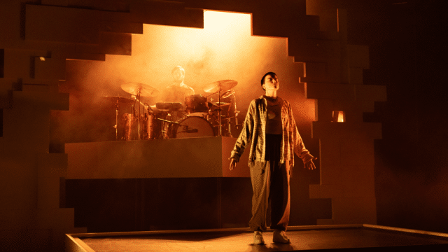 Peak Stuff production photo. A performer wearing wide-legged paper bag trousers and a pastel-coloured flannel shirt stands squarely centre-stage, with their chin titled upwards and their eyes closed, as they are bathed in reddish, hazy light from behind.