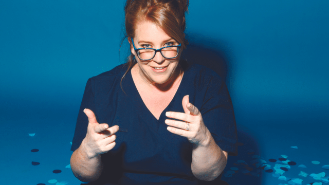 Nurse Georgie Carroll - Sista Flo 2.0 promotional artwork - Georgie Carroll points her index fingers out towards the camera. She is wearing dark blue nurse scrubs and glasses, and she has her hair tied back. The background is blue.