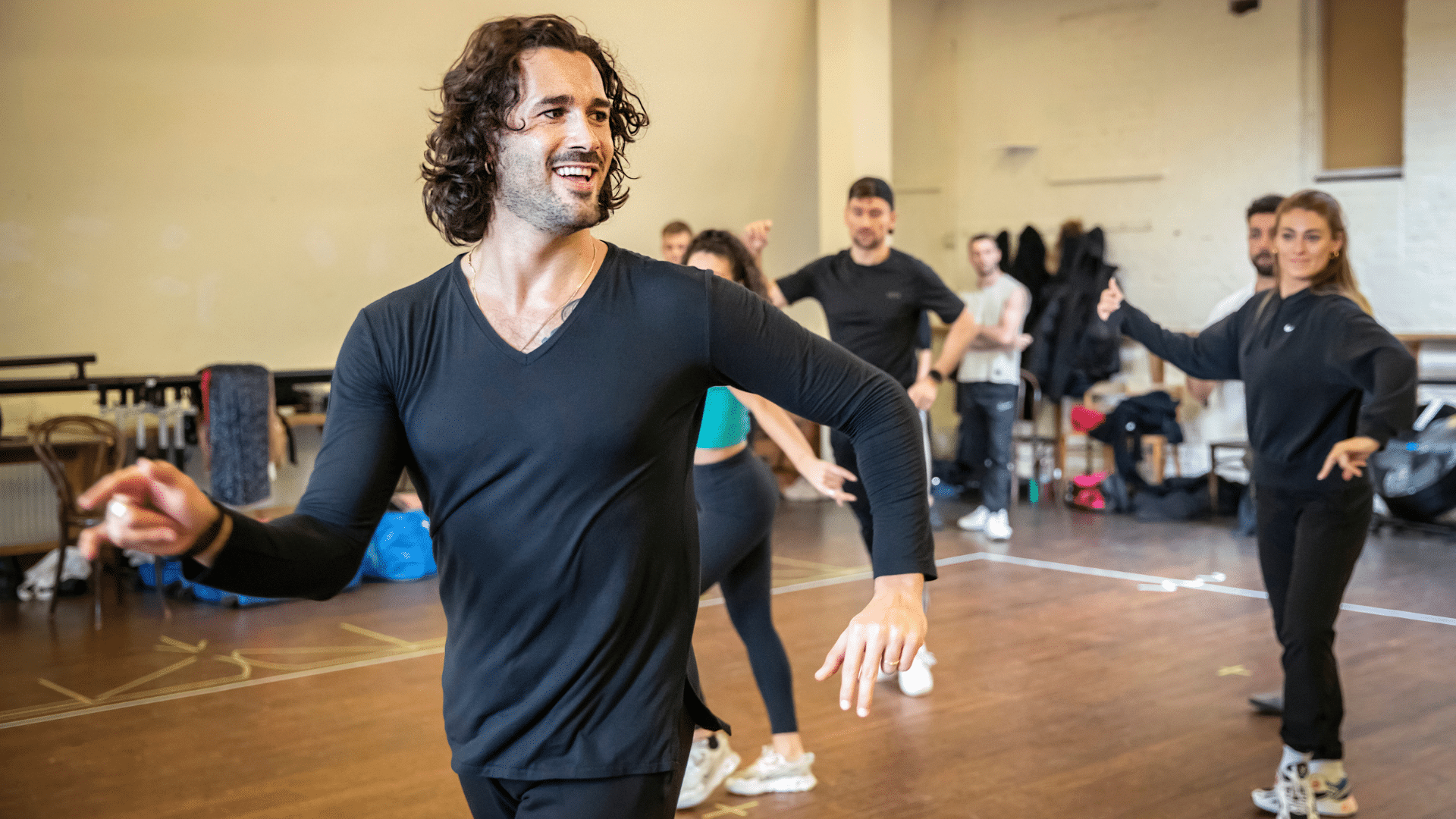Rehearsal photo. Graziano Di Prima seen from the waist up, wearing a black long-sleeved top, jives. He is smiling at a point off-camera. Four other casually-dressed dancers are mimicking his arms position.