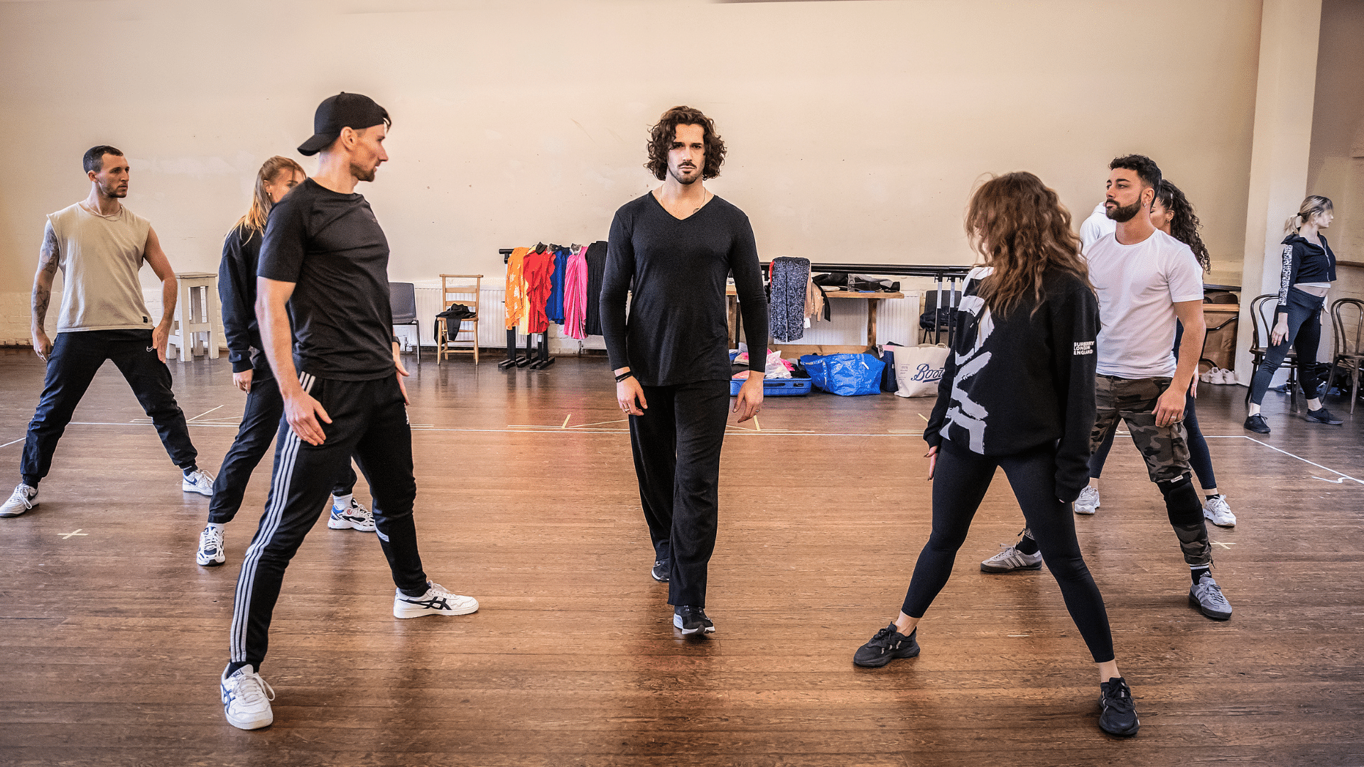 Rehearsal photo. Graziano Di Prima, dressed in all black, walks towards the camera with an intense expression. A triangle of six other dancers stand in a wide-stance and watch him move through their group.