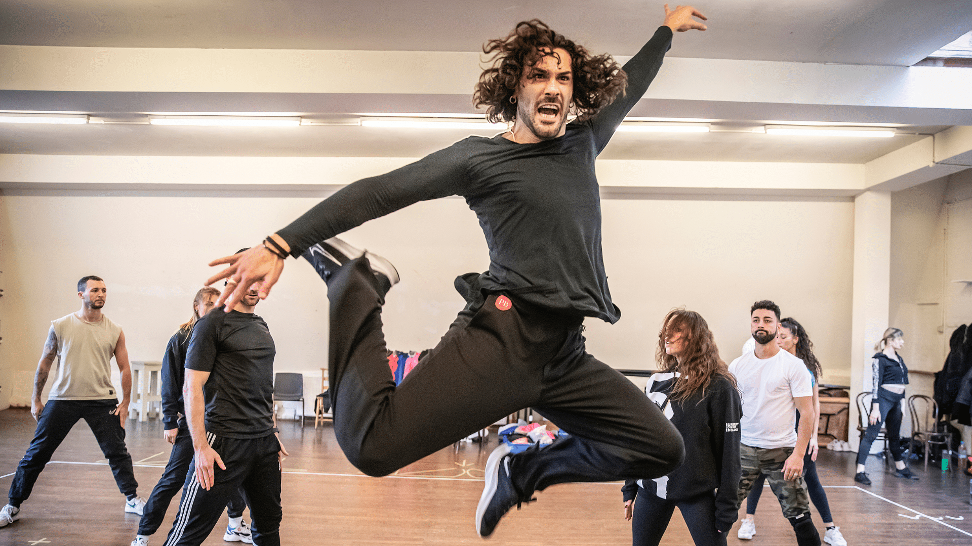 Rehearsal photo. Graziano Di Prima, dressed in all black, performs a balletic leap. His long curly hair is swishing with the movement, his face is contorted with intensity, his arms outspread and tensed. Other dancers look on from behind, apparently standing still.
