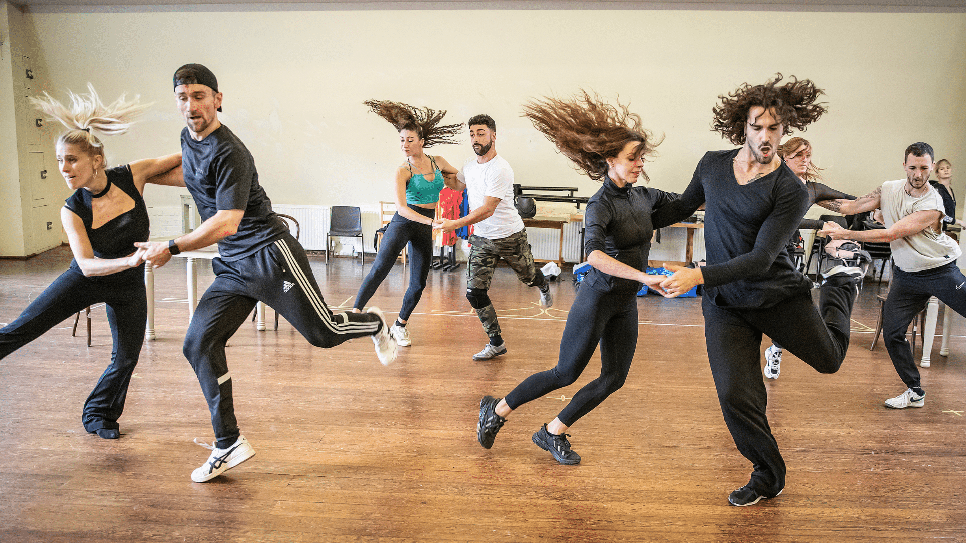 Rehearsal photo. A group of casually-dressed dancers perform in couples, clasping their hands and kicking their outside legs into the air. There is much hair swishing and concentrating faces.