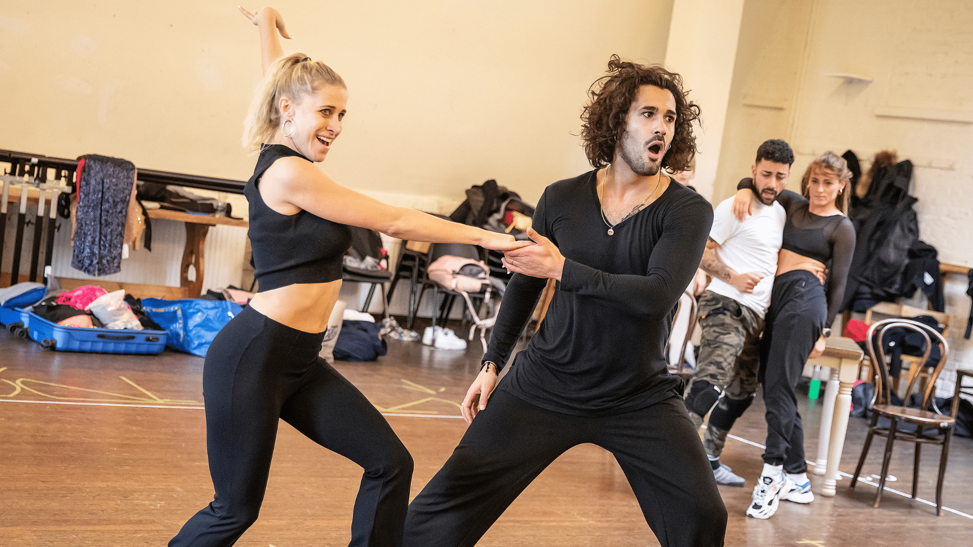 Rehearsal photo. Graziano Di Prima, dressed in all black, clasps hands with a blonde female partner. She is smiling widely, he is gaping open-mouthed.