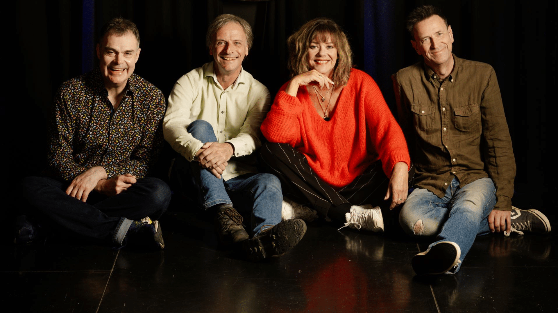 Comedy Store Players Promotional photograph - the four performers sit on the floor of a bare stage, in front of a dark blue theatre curtain. They are all smiling.