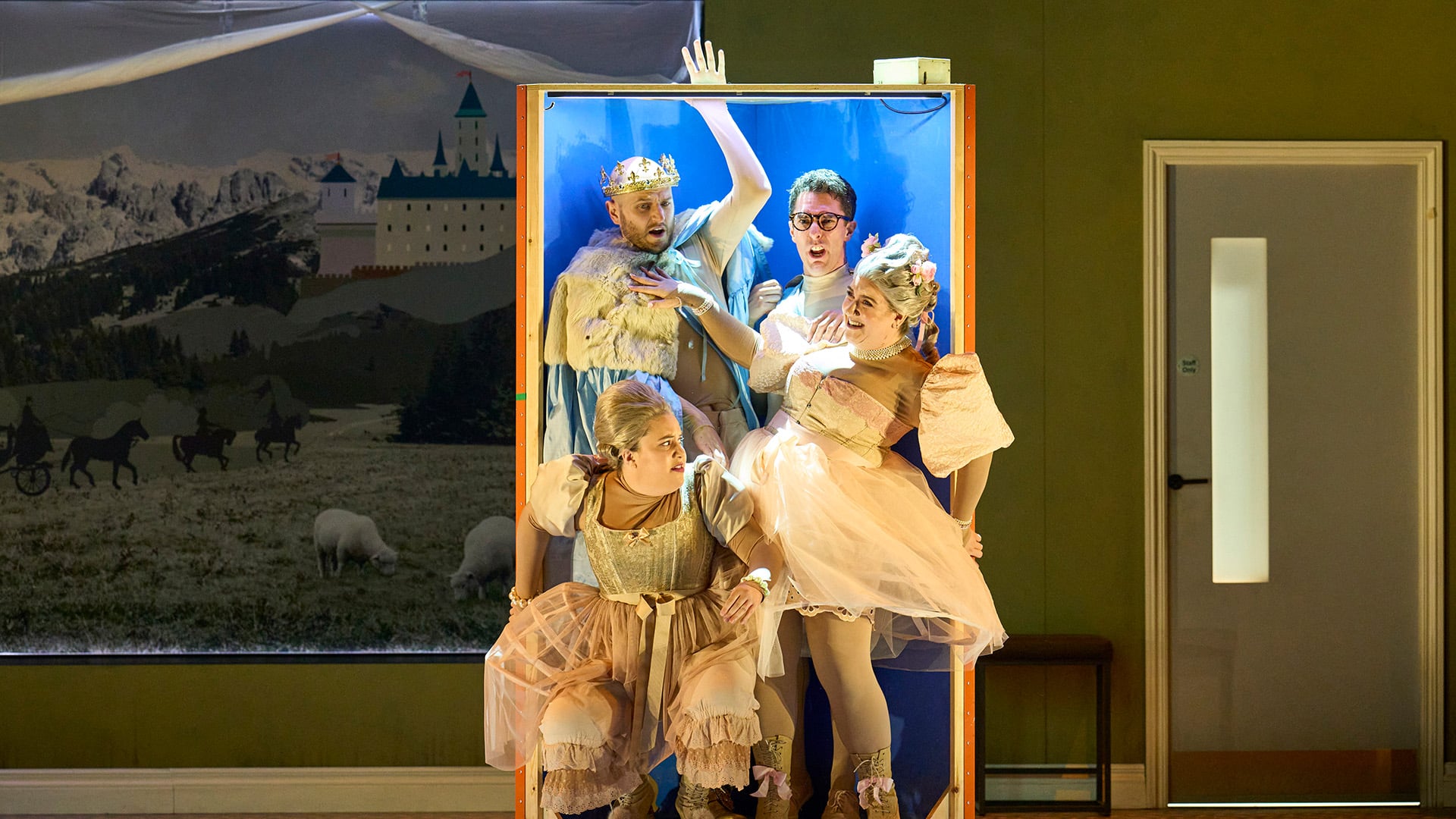 English Touring Opera's Cinderella production photo. A man wearing a golden crown, white fur coat and light-blue fabric looks shocked and raises their arm outside a large wooden box they are stood in. The box has a light blue background. To to right of the man with the crown in the same box, another man wearing a white turtleneck jumper and glasses sneers. Beneath them in the box, two women wearing white frilly Regency dresses laugh and look either side of the box. Behind the group of people in the box, (left) a painting of an alpine country scene with mountains, a castle, and sheep grazing on fields; (right) a lilac door with a sign saying 'Staff Only'.
