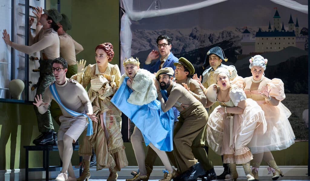 English Touring Opera's Cinderella production photo. A mixed group of people, men wearing beige leotards and Napoleonic army uniforms, and woman wearing white frilly dress, move together as if sneaking around. In the centre of the group, a man wearing a golden grown, white fur coast covering themselves with a large piece of blue fabric. Behind them, beige walls with cabinets filled with swords and a large painting of an alpine castle.