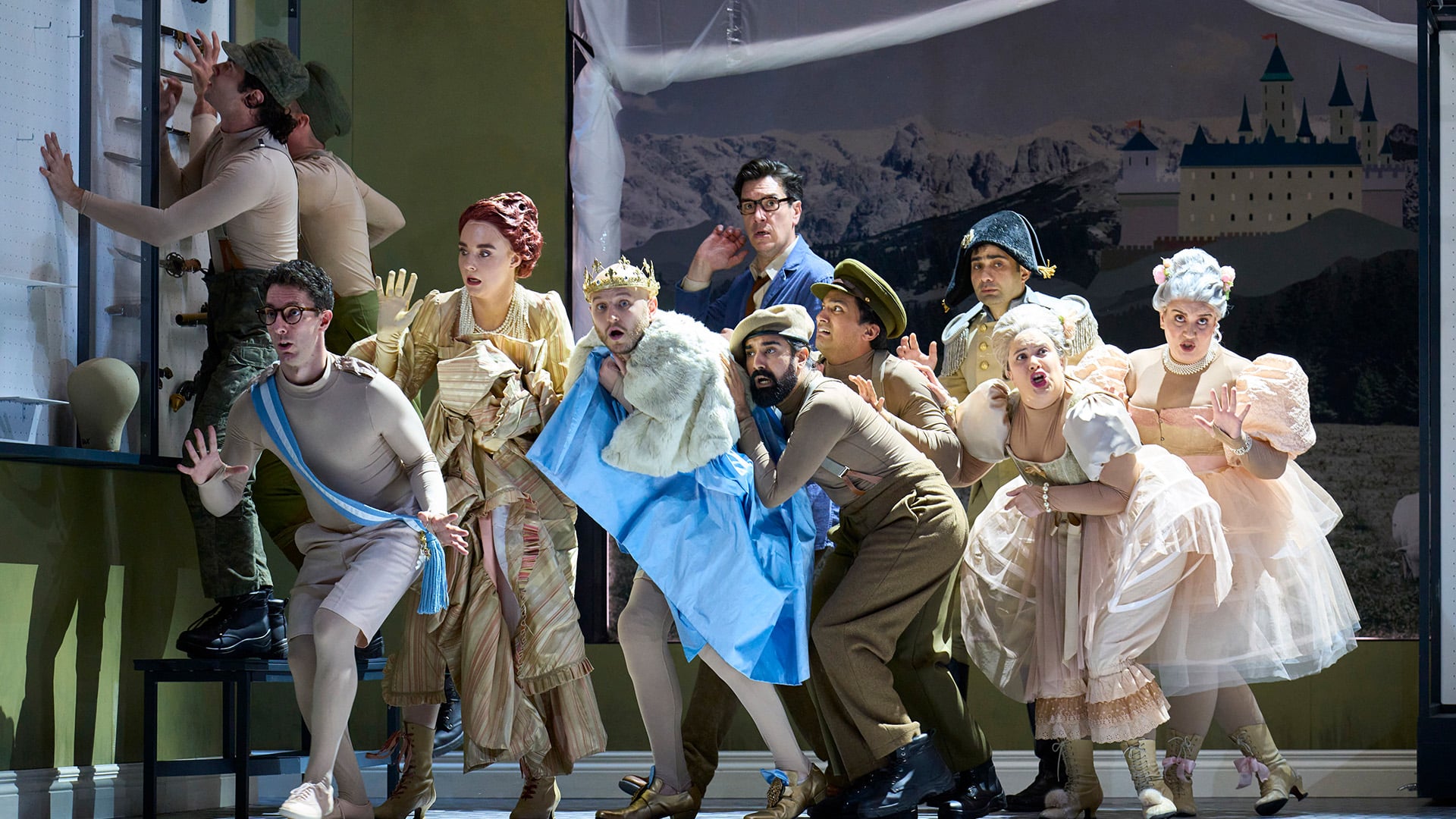 English Touring Opera's Cinderella production photo. A mixed group of people, men wearing beige leotards and Napoleonic army uniforms, and woman wearing white frilly dress, move together as if sneaking around. In the centre of the group, a man wearing a golden grown, white fur coast covering themselves with a large piece of blue fabric. Behind them, beige walls with cabinets filled with swords and a large painting of an alpine castle.
