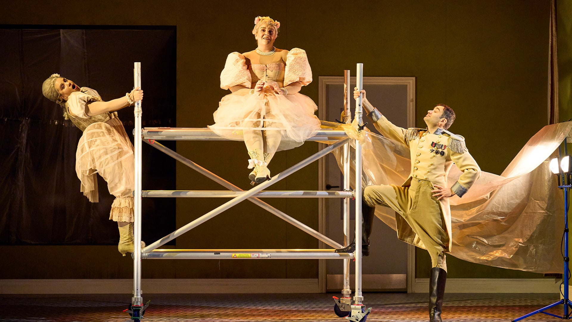 English Touring Opera's Cinderella production photo. A steel scaffolding frame. Left to right: A woman wearing a white frilly dress leans back whilst holding onto the frame; a woman wearing a frilly dress sits on top of the steel frame with their legs cross; a man wearing Napoleonic army officer clothing rests one leg on the steel frame and looks up in accomplishment. Behind the man, a piece of beige tarpaulin hung to look like a flying cape on the man. They are all in a beige-walled room, and there is a lilac door behind them.