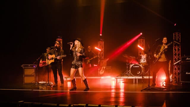 Nashville at Heat: The Modern Country Show production photo. A live band, consisting of two electric guitarists, an acoustic guitarist, a bassist, a drummer and a vocalist, perform on a stage with black flooring and and a black background, lit with red lights.
