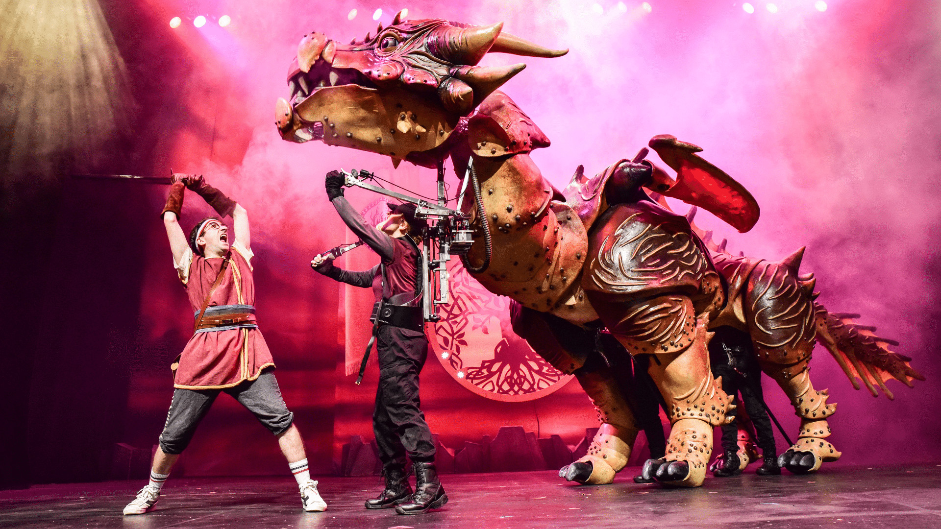 Dragons & Mythical Beasts production photo. Amid smoke and beams of light, a huge, bronze dragon is puppeteered by two performers who are swallowed within its huge mass. A bespectacled man in a red tunic wields a long-sword in the direction of the beast’s ginormous, rearing head.