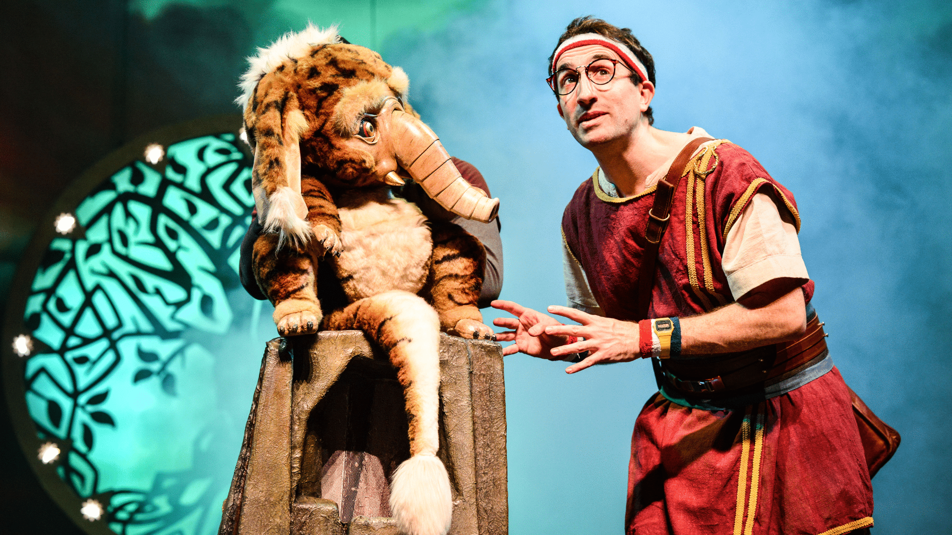 Dragons & Mythical Beasts production photo. A bespectacled man in a red tunic and headband speaks and gestures. Beside him, squatting on a tree stump, is a large, tiger-striped rodent with floppy ears, and a small trunk and tusks. It listens to the man with warm, intelligent eyes.