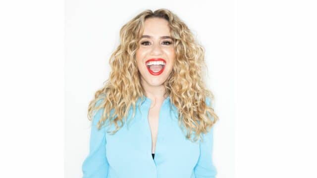 Comedian Michelle Brasier is wearing a bright blue shirt, her hair is down in loose curls and she's wearing a bright red lipstick. She's facing the camera with a big, bright, toothy smile.