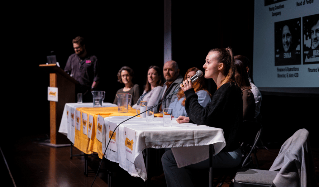 A photograph from Exeter Northcott's Speakers for Schools event: a young woman sitting in a panel holds a microphone and smiles.