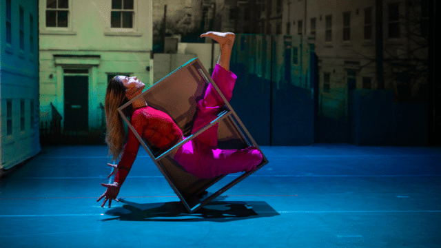 Nobody production image: a woman in a fushia top and trousers dancing while enclosed in a small metal cube, which balances on its edge; her arms and legs are flexed and reaching out of the cube; the background is a grey urban landscape.