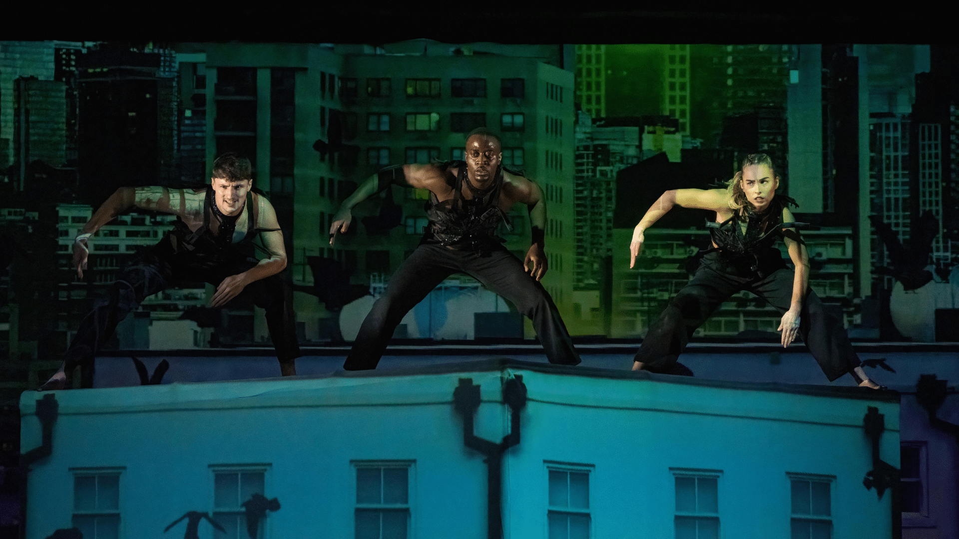 Nobody production photo: three performers dressed in black crouch side-by-side on a platform in an urban landscape on stage.