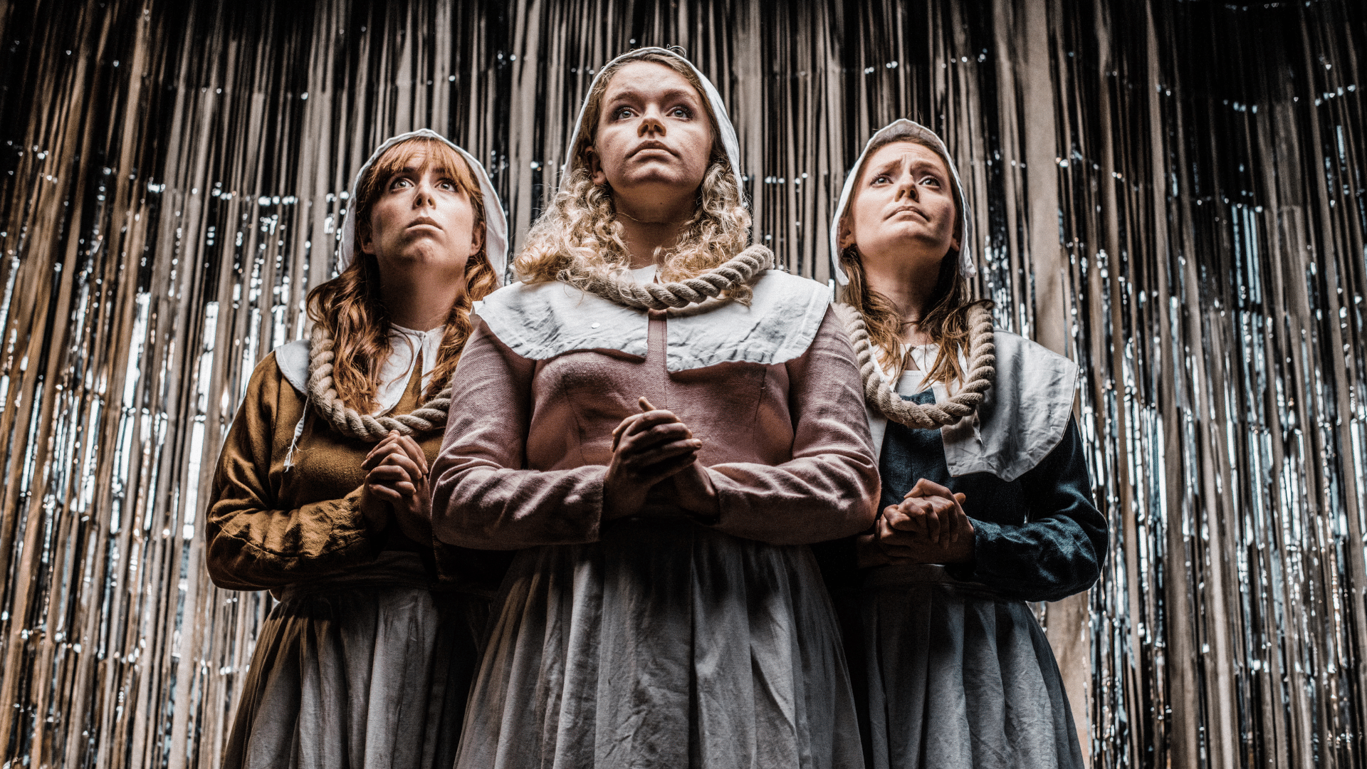 Hags promo photo. A low-angle shot, looking up at three performers in medieval puritan dress, clasping their hands in prayer and looking to the heavens with pious expressions. The background is a silver glittery curtain.