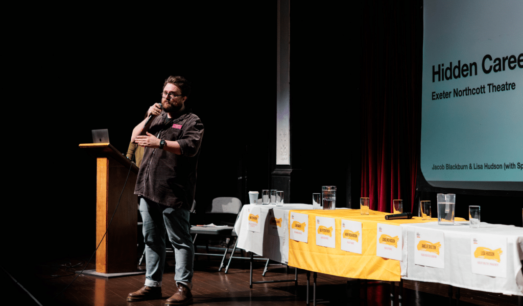A photograph from the Northcott's Speakers for Schools event: Jacob Blackburn, wearing jeans and a dark shirt, stands on stage and gestures.