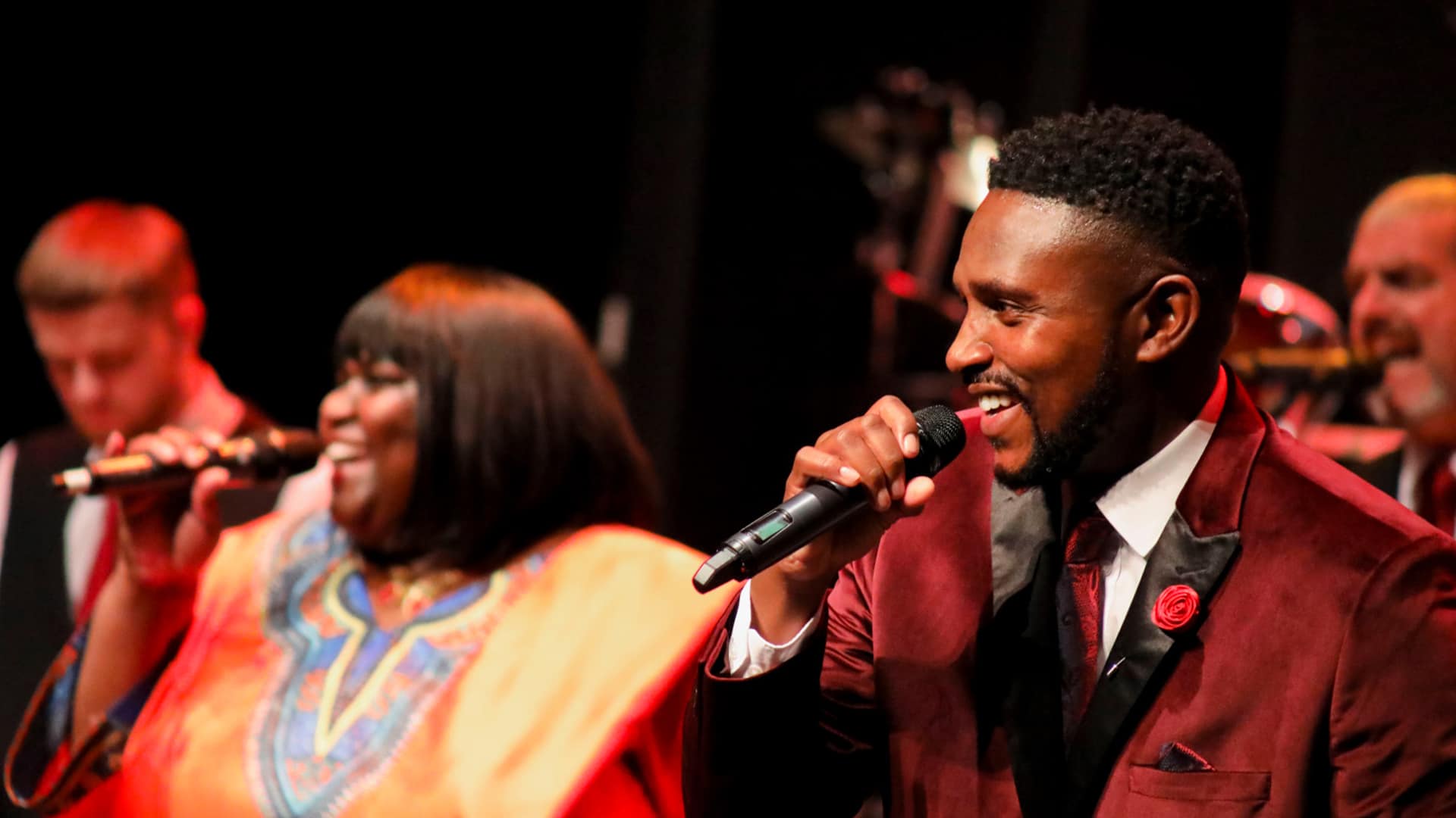 RUSH: A Joyous Jamaican Journey production photo. (Left to right) A black woman wearing an orange dress with a blue and light orange patten down the centre and a black man wearing a red velvet jacket over a white shirt and black tie sing into microphones.