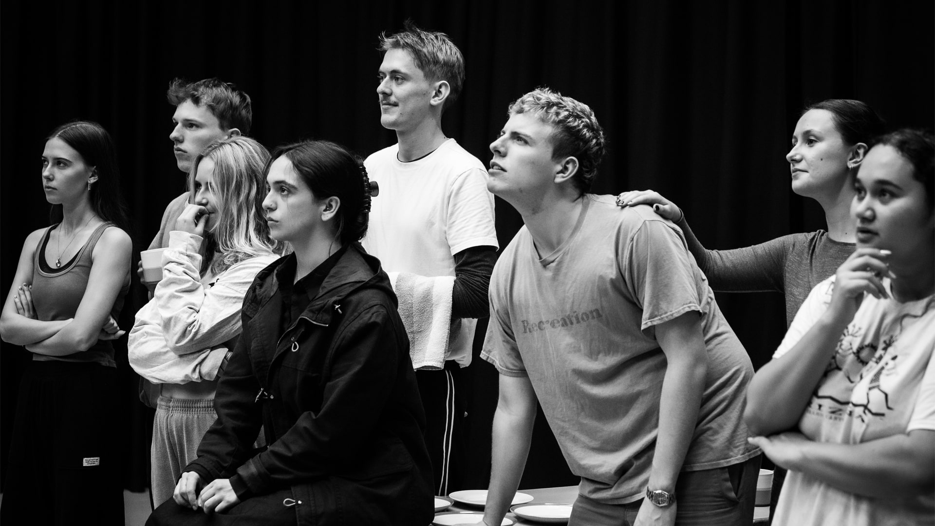 Alice rehearsal photo: monochrome; eight casually-dressed young people gather around a table laid with china plates, their attention focused on a point off-camera to the left.