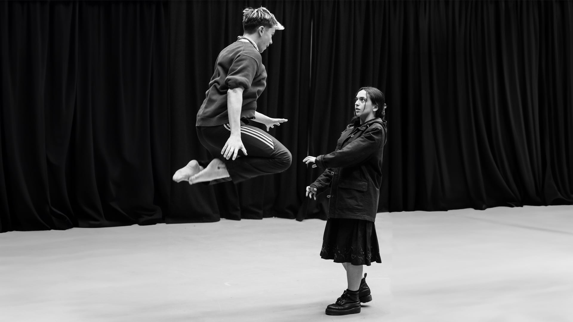 Alice rehearsal photo: monochrome; a young person dressed in a black midi-skirt and long dark jacket looks shocked as another young actor, wearing a striped tracksuit, jumps into the air in front of her.