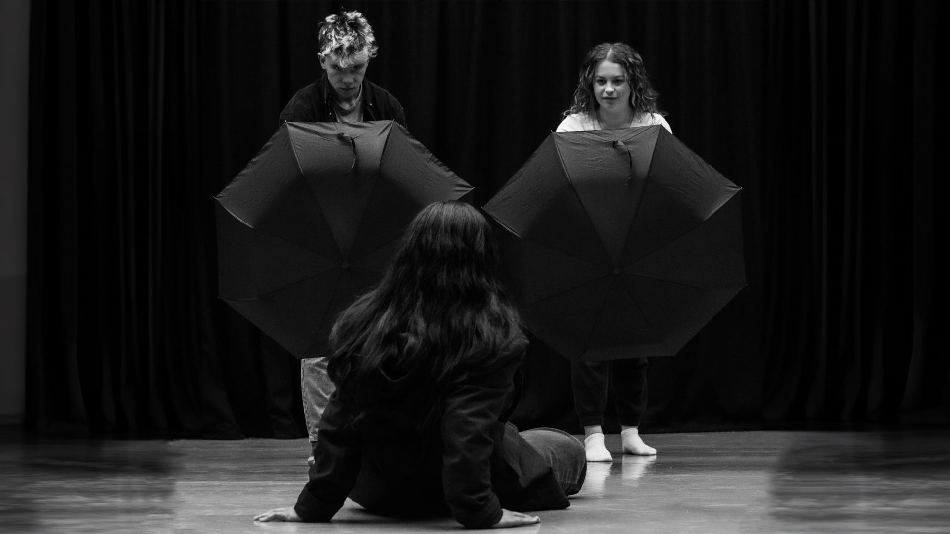 Alice Rehearsal Photo: monochrome; an actor with long dark sits on the floor with her hands braced against it behind her, facing away from the camera; two other young actors tower over her, each holding an opened umbrella like a shield towards the camera.