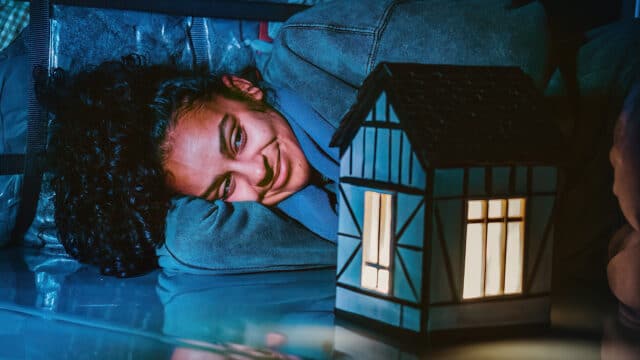 Belongings artwork – a young woman with curly brown hair wearing a blue jumper lies in a foetal position smiling at a small sculpture of a house on a mirrored floor.