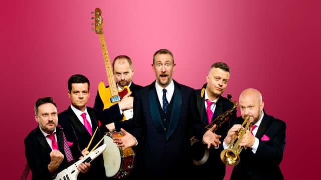 The Horne Section's Hit Show! artwork. Pink gradient background. Members of The Horne Section stand together, with creator Alex Horne in the centre, wielding different musical instruments. They are all wearing matching Navy suits.