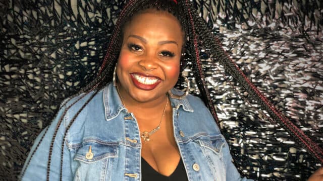Jackie Fabulous photo. Jackie Fabulous, a black woman with long brown braided hair, wearing a denim shirt over a black top, smiling. She is stood in front of tinsel covered wall.