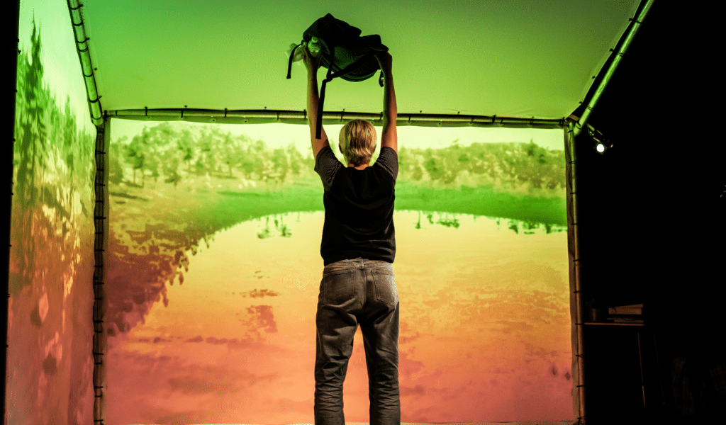 Feel Me production photo. A performer stands facing away from the camera with a black rucksack raised above their head. Behind them, a cube made of gauze which can be projected onto. It is missing two side walls, and the back walls are displaying a brightly coloured and highly-saturated photograph of a stagnant body of water, bordered by short, scrubby trees.