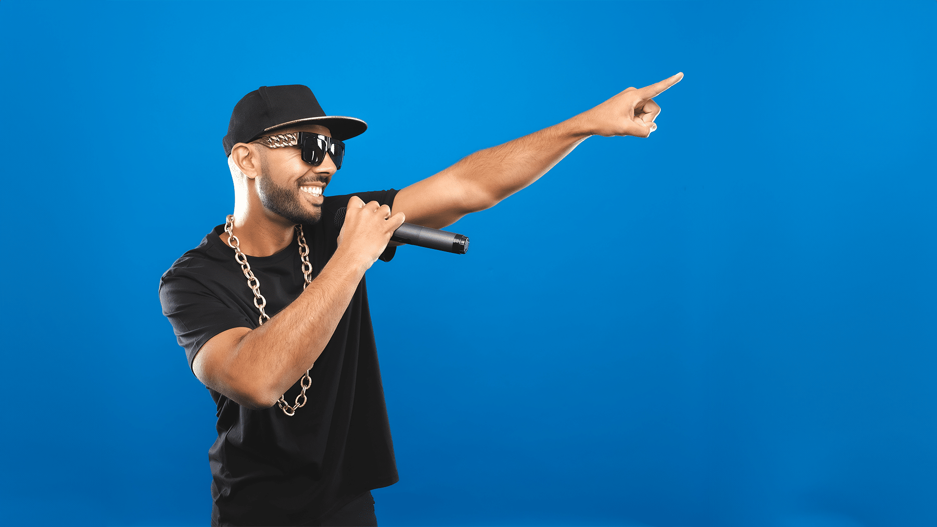 MC Grammar promotional artwork. Blue background. MC Grammar holds a microphone to his smile and points off-camera, wearing a black t-shirt, gold chain necklace and black baseball cap.