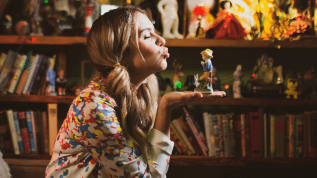 Background: a bookshelf lit by fairy lights and crammed with models, toys, and books. Michele Brasier, wearing her long blonde hair in two ponytails either side of her face, is seen in profile. She is holding a painted miniature boy in her flat palm, towards whom she is puckering.