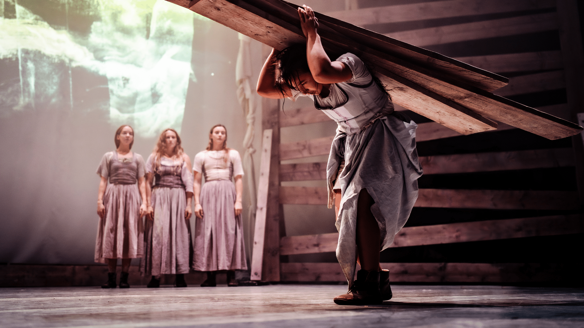 Tess production photo: a performer in a full-skirted, pale linen dress struggles to carry 4 huge planks of wood on her shoulders; in the background, three other woman, similarly dressed, look on.