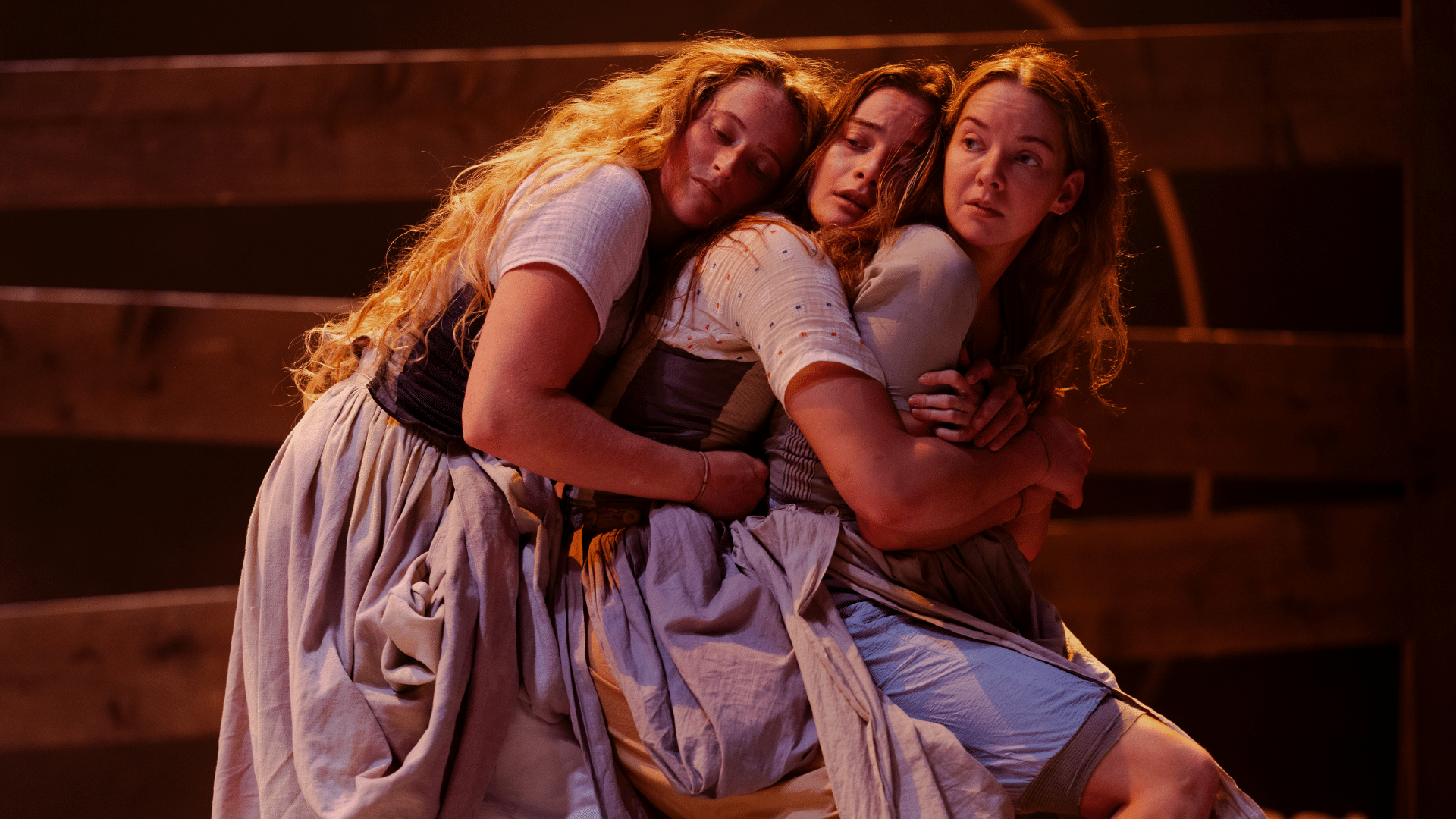 Tess production photo: three performers dressed in similar, pale, linen dresses hug one another from behind; they are bathed in a warm glow.