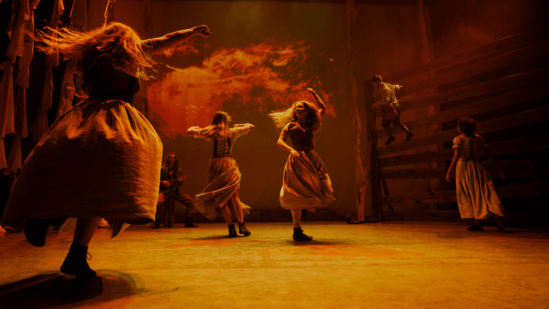 Tess production photo: four performers twirl in full-skirted white dresses, bathed in an orange glow; on the wall behind them, a red fire is projected.